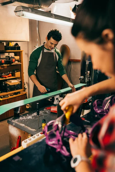 Selective focus of smiling worker repairing ski and colleague on background — Stock Photo