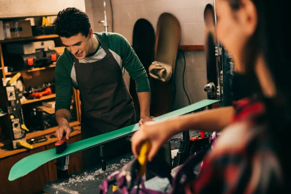 Selektiver Fokus des lächelnden Arbeiters, der Ski mit Bürste und Kollege im Hintergrund putzt — Stockfoto