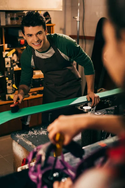 Selektiver Fokus des lächelnden Arbeiters, der Ski mit Bürste und Kollege im Hintergrund putzt — Stockfoto