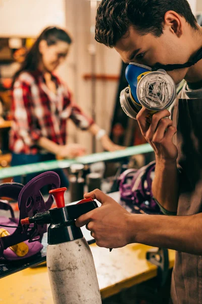 Selective focus of worker in respirator spraying on snowboard in repair shop — Stock Photo