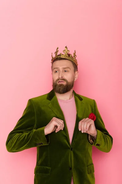 Hombre guapo con corona en chaqueta de terciopelo mirando hacia otro lado sobre fondo rosa - foto de stock