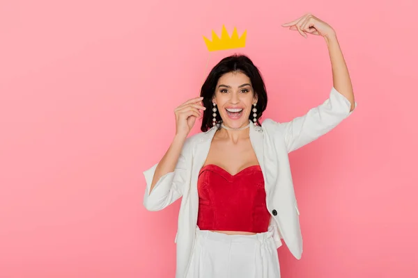 Smiling woman pointing with finger at paper crown on pink background — Stock Photo