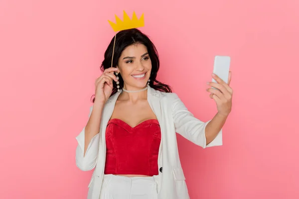 Mulher sorrindo segurando coroa de papel e tomando selfie no fundo rosa — Fotografia de Stock