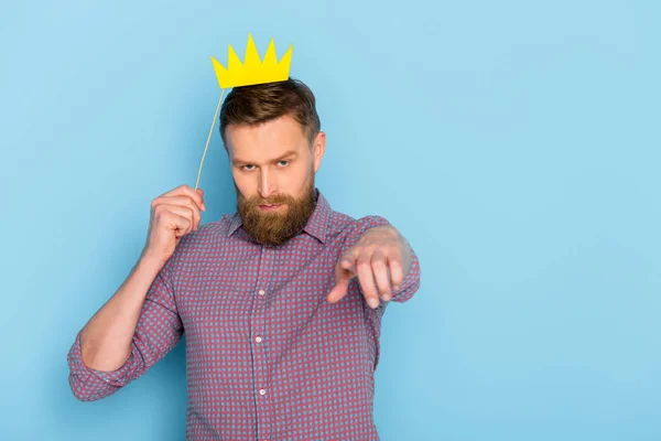 Serious man holding paper crown and pointing with finger on blue background — Stock Photo