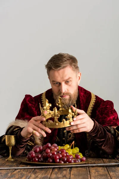 Rey sentado a la mesa y sosteniendo la corona aislada en gris - foto de stock