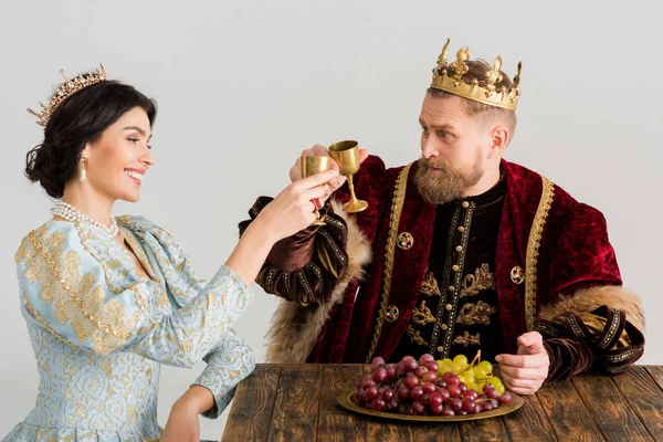 Smiling queen and king with crowns clinking with cups isolated on grey — Stock Photo