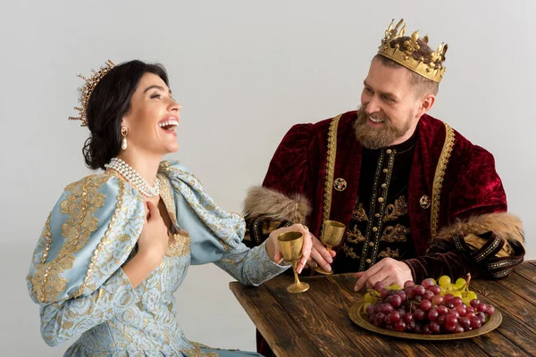 Smiling queen and king with crowns sitting at table isolated on grey — Stock Photo