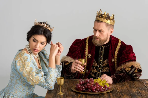 Upset queen and king with crowns sitting at table isolated on grey — Stock Photo