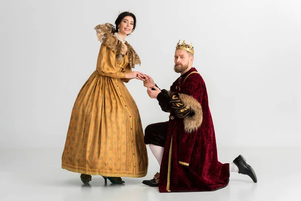 King with crown bending on knee and holding hand of smiling queen on grey background — Stock Photo