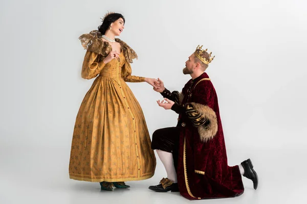 King with crown bending on knee and holding hand of shocked queen on grey background — Stock Photo