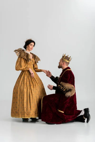 King with crown bending on knee and holding hand of queen on grey background — Stock Photo