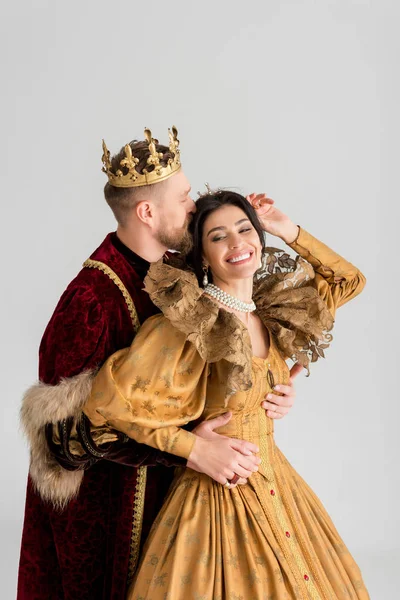 Король с короной целует и обнимает улыбающуюся королеву, изолированную на серой — стоковое фото