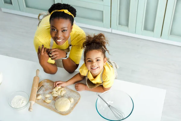 Top view of cheerful african american mother and daughter looking at camera while sculpting dumplings — Stock Photo