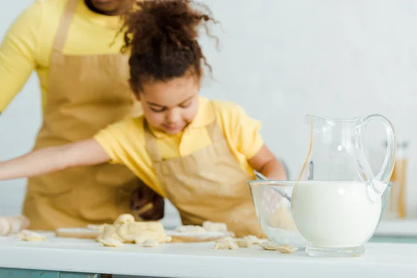 Focus selettivo di brocca con latte vicino ciotola e carino bambino africano americano scolpire gnocchi vicino alla madre — Foto stock