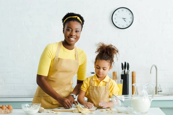 Glücklich afrikanisch-amerikanische Mutter und süße Tochter in Schürze Modellierung rohe Knödel in der Küche — Stockfoto