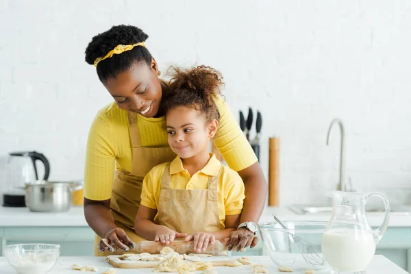 Mignon afro-américain enfant tenant rouleau à pâtisserie près de mère gaie et des boulettes crues sur planche à découper — Photo de stock