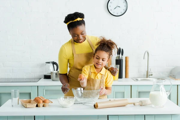 Cute african american kid holding egg near cheerful mother and bowl — Stock Photo