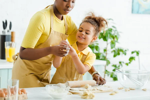 Enfoque selectivo de niño afroamericano lindo sosteniendo masa cruda cerca de la madre y albóndigas en la tabla de cortar - foto de stock