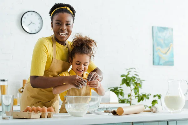 Lindo africano americano niño y alegre madre añadir huevo en bowl mientras cocinar en cocina - foto de stock