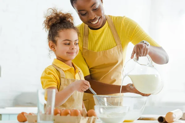 Selective focus of happy african american mother pouring milk in bowl near cute daughter — Stock Photo