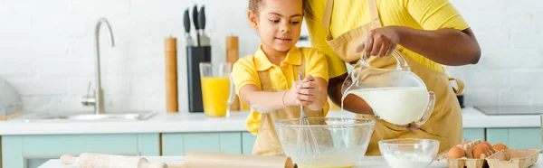 Panoramic shot of african american mother pouring milk in bowl near cute daughter — Stock Photo