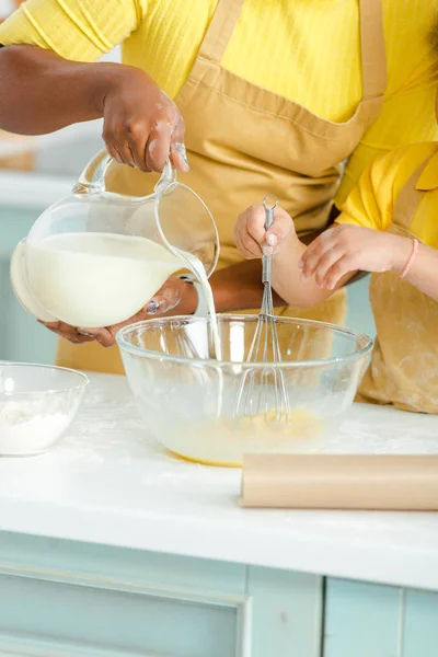 Vista recortada de la madre afroamericana vertiendo leche en un tazón cerca de su hija - foto de stock