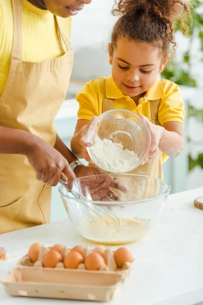 Selektiver Fokus der süßen afrikanisch-amerikanischen Kind hält Schüssel mit Mehl in der Nähe der Mutter in der Küche — Stockfoto