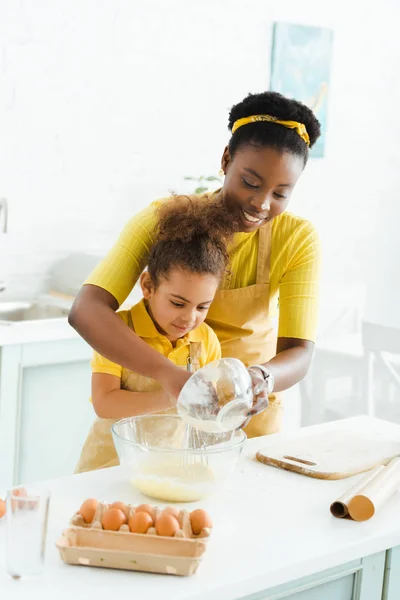 Glücklich afrikanisch-amerikanische Mutter hält Schüssel mit Mehl in der Nähe niedliche Tochter in der Küche — Stockfoto