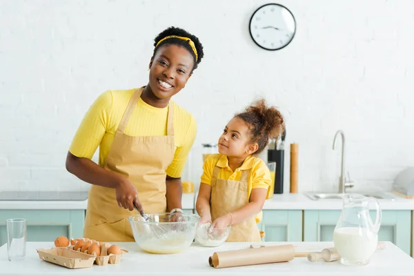 Cute african american kid looking at happy mother while putting hands in bowl with flour — Stock Photo