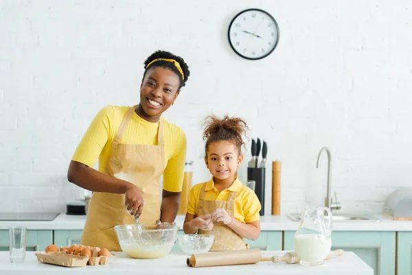 Niño afroamericano feliz y madre alegre sonriendo mientras cocina en la cocina - foto de stock