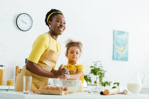 African american kid and cheerful mother smiling while cooking in kitchen — Stock Photo