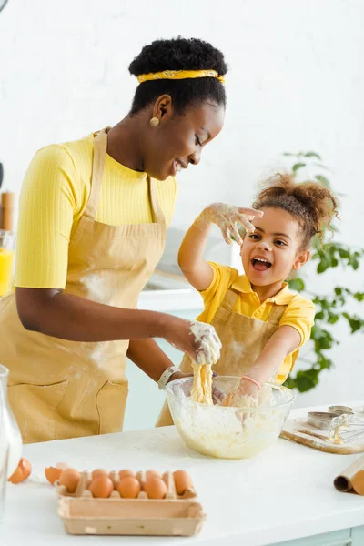 Счастливая африканская американская мать месит тесто рядом с веселой дочерью — стоковое фото