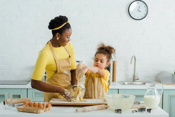 Mãe americana africana alegre e filha amassar massa de farinha perto de ingredientes na cozinha — Fotografia de Stock