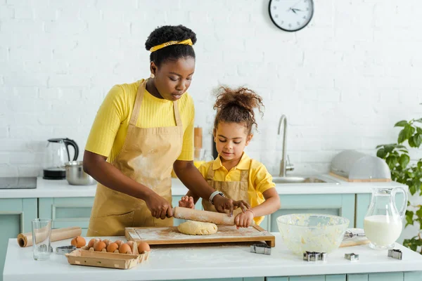 Carino africano americano figlia guardando grezzo pasta vicino madre holding mattarello — Foto stock
