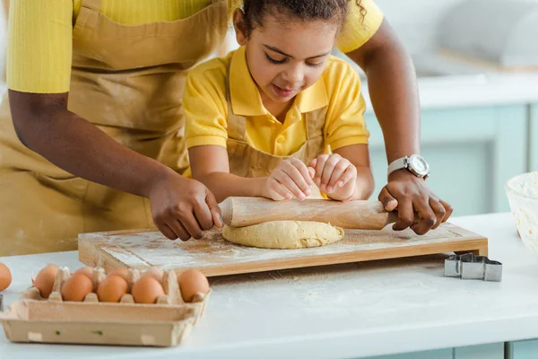Обрезанный вид на африканскую американскую мать, держащую скалку рядом с тестом и симпатичную дочь на кухне — стоковое фото