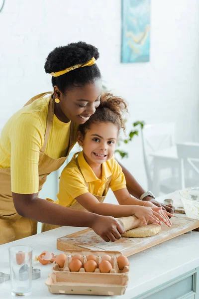 Attractive african american mother looking at raw dough near cute daughter and holding rolling pin — Stock Photo