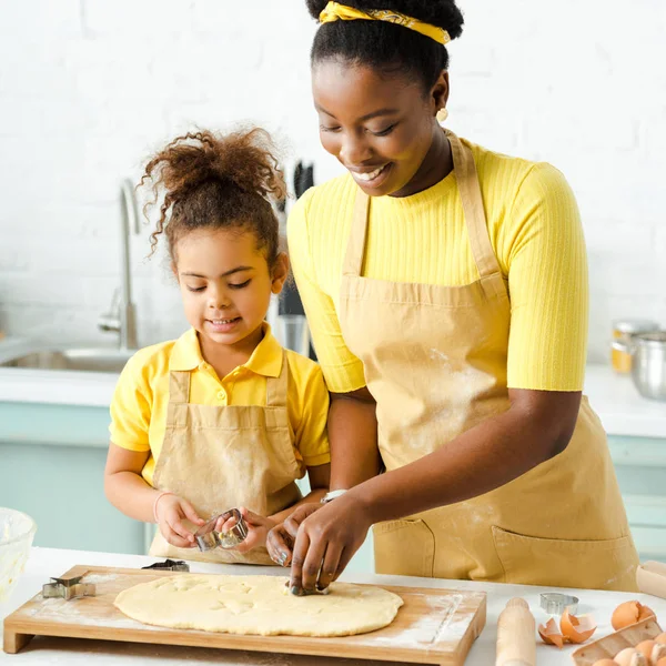 Adorável criança afro-americana e feliz mãe segurando cortadores de biscoitos perto da massa crua — Fotografia de Stock