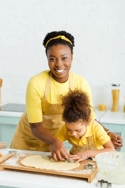 Criança americana africana feliz e mãe alegre segurando cortadores de biscoitos perto da massa crua na cozinha — Fotografia de Stock