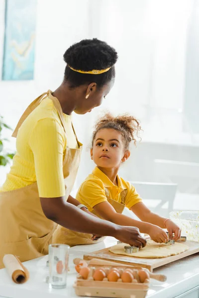 Süße afrikanisch-amerikanische Kind schaut Mutter beim Halten von Ausstechformen in der Nähe rohen Teigs in der Küche — Stockfoto