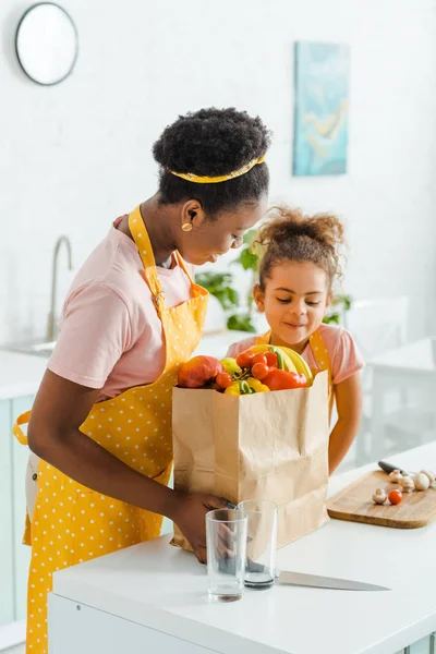 Atractiva afroamericana madre e hija mirando bolsa de papel con comestibles - foto de stock