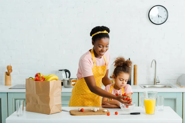 Feliz africano americano madre poniendo tomates cherry en tazón cerca de hija - foto de stock