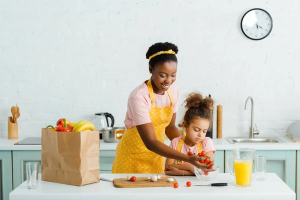 Happy african american mother putting cherry tomatoes in bowl near daughter — Stock Photo