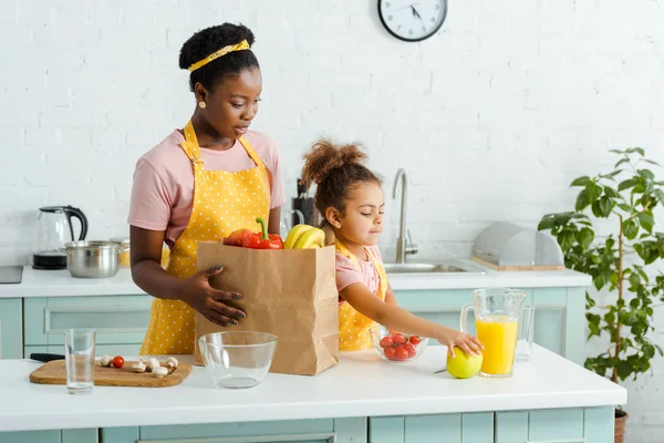 Африканская американская мать смотрит на дочь, берущую яблоко на кухне — стоковое фото