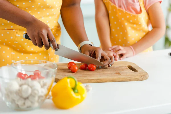 Cropped view of african american woman cutting cherry tomatoes on cutting board near daughter — Stock Photo