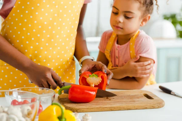 Vue recadrée de femme afro-américaine coupant poivron sur planche à découper près de fille mignonne — Photo de stock