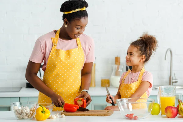 Happy african american woman cutting bell pepper on cutting board near cute daughter — Stock Photo