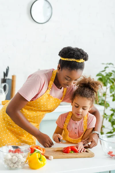 Happy african american woman cutting bell pepper with cute daughter — Stock Photo