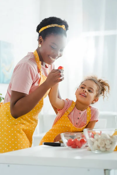 Selective focus of happy african american mother holding cherry tomato near kid — Stock Photo