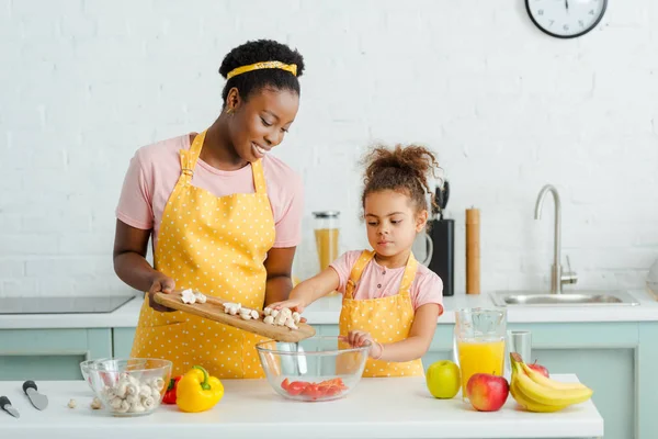 Cheerful african american mother holding cutting board with mushrooms near bowl and  cute daughter — Stock Photo