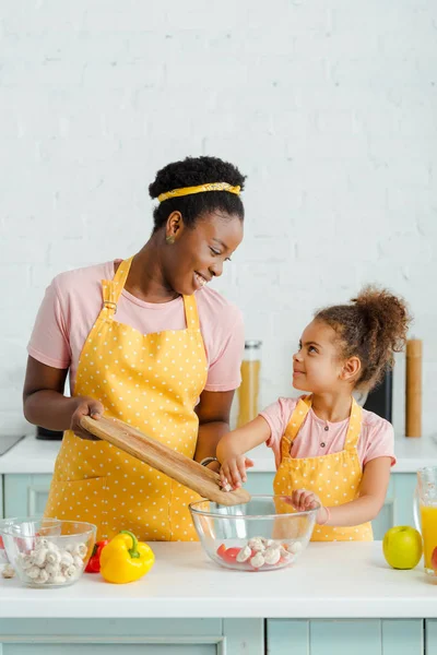 Cheerful african american mother holding cutting board near bowl and looking at cute daughter — Stock Photo